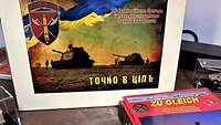Auf einem braunen Tisch liegen rote Bücher, ein buntes ukrainisches Bild und Panzermodelle.