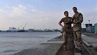 Zwei deutsche Soldaten stehen auf einer Pier und besprechen sich. Im Hintergrund stehen mehrere Schiffe.
