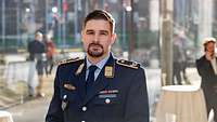 Major Robin N. Auf dem Porträtbild blickt Major Robin N. in die Kamera. Hinter ihm sind Stehtische aufgebaut