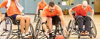 Drei Sportler im Rollstuhl kämpfen um einen auf dem Boden rollenden Basketball.