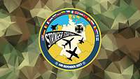 Vor einem grünen Flecktarn-Polygon-Hintergrund ist das runde Patch von Air Defender 2023.