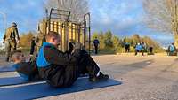 Soldaten machen um einen gelben Container herum Fitnessübungen in schwarz-blauen Trainingsanzügen.