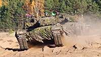 Drei Panzer fahren hintereinander auf sandigem Boden. Im Hintergrund bewaldetes Gelände. 