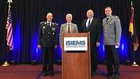 Four men standing on the podium, two of them in uniform, the German and American flag on either side