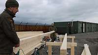 Auf einer Baustelle stehen zwei Soldaten vor einen Stapel Holzbalken, Die beiden Soldaten halten dabei einen Balken hoch.