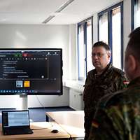 Zwei Soldaten reden miteinander, im Hintergrund ein Laptop und ein großer Bildschirm.