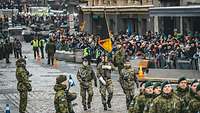 Drei Soldaten der Luftwaffe marschieren mit einer Truppenfahne über einen Marktplatz, umgeben von Zuschauern und Soldaten.