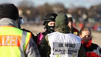 Ein Bundeswehrarzt in Flecktarn mit Einsatzweste darüber spricht mit einem Polizisten.