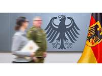 Ein Soldat und eine Zivilistin (mit einer Urkunde in der Hand) vor einer Wand mit dem Bundesadler