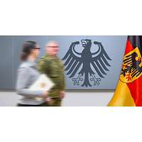Ein Soldat und eine Zivilistin (mit einer Urkunde in der Hand) vor einer Wand mit dem Bundesadler