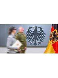 Ein Soldat und eine Zivilistin (mit einer Urkunde in der Hand) vor einer Wand mit dem Bundesadler