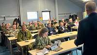 Soldiers from different nations sit at tables and look ahead at a screen.