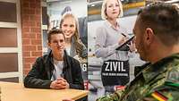 Blick über die Schulter eines Soldaten. Er spricht mit einem jungen Mann. Dahinter zwei Plakate. 