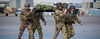 Soldaten aus mehreren Nationen haben einen Verwundeten auf einer Trage und marschieren entlang eines Flugfelds. 