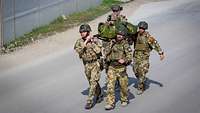Fünf Soldaten tragen einen Verwundeten auf einer Trage eine Straße entlang.