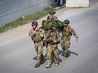 Fünf Soldaten tragen einen Verwundeten auf einer Trage eine Straße entlang.