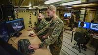 Zwei Soldaten arbeiten im Stehen an einem Computer, auf dem Luftbildaufnahmen zu sehen sind.