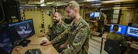 Zwei Soldaten arbeiten im Stehen an einem Computer, auf dem Luftbildaufnahmen zu sehen sind.