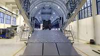 Simulator in Form eines Flugzeugrumpfes mit geöffneter Laderampe in einer Halle.