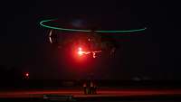 In der Nacht schwebt ein Hubschrauber, der beleuchtet ist, nah über eine Bodencrew, die am Boden steht