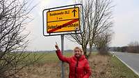 Eine Frau im roten Mantel zeigt auf ein gelbes Ortsschild am Rande der Stadt