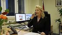 Eine Frau im schwarzen Kostüm mit blonden Haaren sitzt am Schreibtisch in einem Büro und telefoniert
