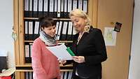 Zwei Frauen stehen in einem Büro vor einem Aktenschrank und lesen in einem Papierdokument 