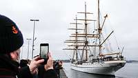 Ein weißes, dreimastiges Segelschiff liegt ohne gesetzte Segel neben einer Hafenpier.