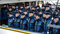 Marinesoldaten in blauer Arbeitsjacke stehen in drei Reihen angetreten.