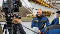 Ein Marineoffizier in blauer Arbeitsjacke seht vor einer Videokamera.