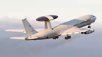 Ein Flugzeug vom Typ AWACS fliegt vom Betrachter weg, im Hintergrund sind Wolken zu erkennen.