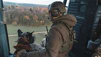 Ein Hund und ein Soldat sitzen nebeneinander an der offenen Tür eines Hubschraubers beim Flug.