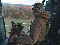 Ein Hund und ein Soldat sitzen nebeneinander an der offenen Tür eines Hubschraubers beim Flug.