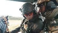 Zwei Soldaten sitzen in einem Hubschrauber und unterhalten sich. Einer hält einen Hund an der Leine.