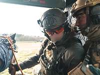 Zwei Soldaten sitzen in einem Hubschrauber und unterhalten sich. Einer hält einen Hund an der Leine.