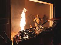 Drei Soldaten laufen mit einem Hund durch ein Gebäude in dem ein Feuer brennt und ein Autowack steht.