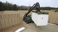 Ein militärischer Containerstapler fährt zwischen Hesco-Wänden mit einem weißen Container am Ladearm. 