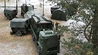 Militärfahrzeuge befinden sich unter Bäumen bei leichtem Schneefall. Sie werden von einem Soldaten eingewiesen. 