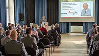 Ein Soldat spricht zu den im Saal sitzenden Soldatinnen und Soldaten sowie Zivilisten.