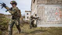 Ein bewaffneter Soldat läuft von einer Häuserwand weg, an der kniet ein anderer Soldat und sichert.