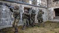 Vier Soldaten in Flecktarnuniform stehen, jeweils mit einem Gewehr bewaffnet, in einer Häuserruine.
