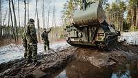 Drei Soldaten stehen vor einem Pionierpanzer Dachs auf einem schlammigen Waldweg