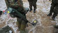 Ein Soldat hält den Dual-Sensor-Detektor VMR3 in der Hand und führt ihn über den schlammigen Boden.
