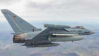Ein Eurofighter fliegt über eine Landschaft. Am Boden sieht man Felder und Häuser.