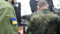 Eine Person trägt ein ukrainisches Flaggen-Patch am Oberarm. Im Hintergrund ist unscharf ein deutscher Soldat zu sehen.
