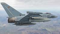 Ein Eurofighter fliegt über eine Landschaft. Am Boden sieht man Felder und Häuser.