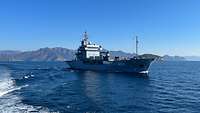 Ein Hilfsschiff der Typ-Klasse Tender auf offenem Meer. Im Hintergrund sind Berge zu sehen.