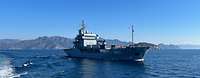Ein Hilfsschiff der Typ-Klasse Tender auf offenem Meer. Im Hintergrund sind Berge zu sehen.
