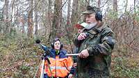 Umweltschutzmessung Schießbefehl Ein Mann in Schutzjacke und ein Mann in Uniform stehen in einem Waldstück mit technischen Geräten.