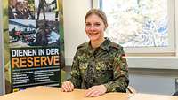 Eine Soldatin sitzt lächelnd an einem Tisch, dahinter ein Roll-up „Dienen in der Reserve“
